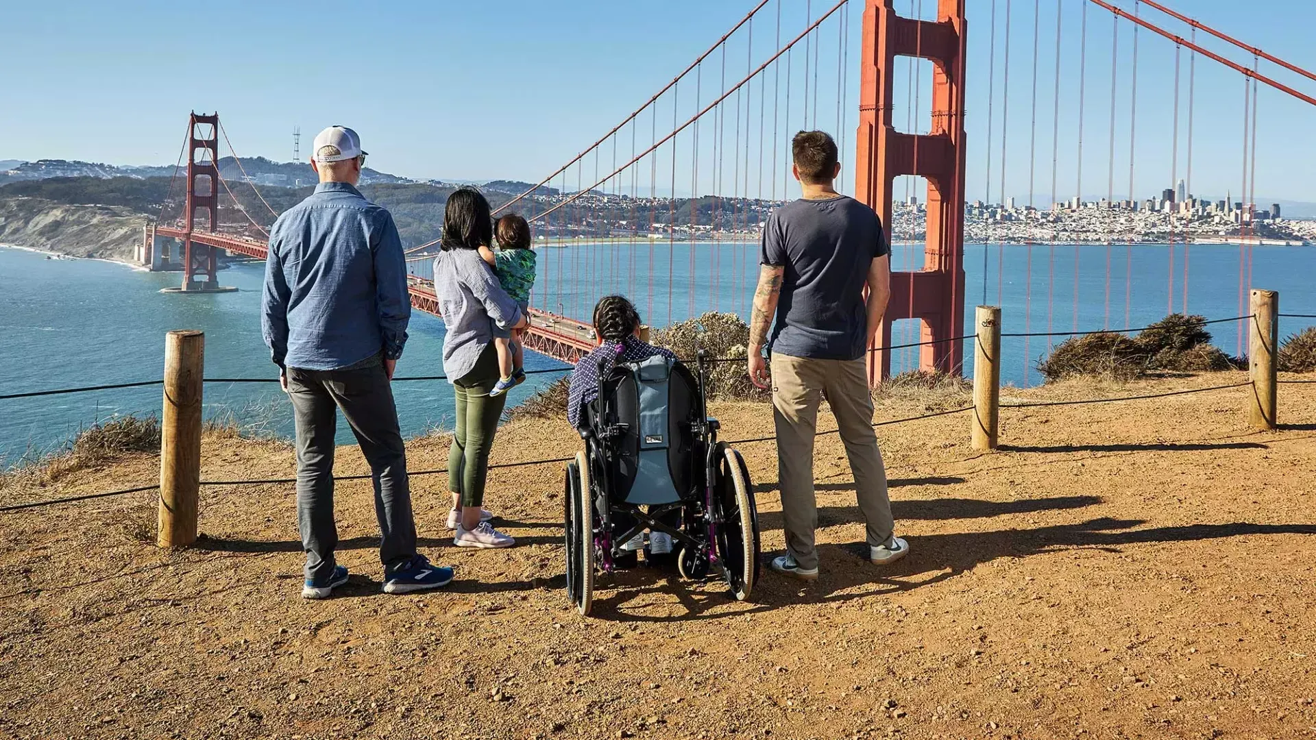 Eine Gruppe von Menschen, 包括一个坐轮椅的人, ist von hinten zu sehen, während sie von den Marin Headlands aus auf die Golden Gate Bridge blicken.