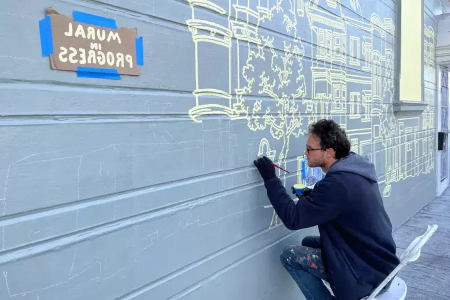 一位艺术家在教会区一栋建筑的侧面画壁画, 大楼上贴着一块牌子，上面写着"壁画正在进行中". São Francisco, Califórnia.